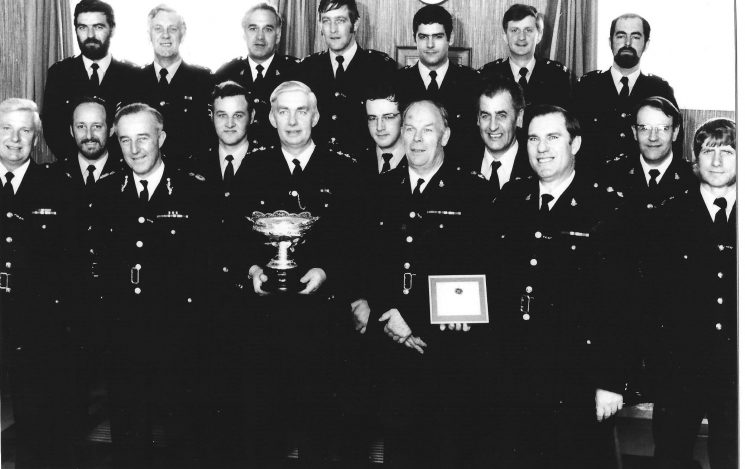 Officers of Cirencester Sub Division at presentation of Caroline Symes Memorial Bowl for Community Service 1978. Back row left to right - Police Constable P. Clare; Police Sergeant H. Eastwood;  Inspector WF. Jones; Police Constables M. Lees; C. Heselton (under the clock);  B. Manning; D. Legg. Middle row - Police Constables E. Heath; J. Squires; D. Causon;  Police Sergeant D. Smeeton;  Inspector V. Causon. Front row - Superintendent M. Cooper;  Chief Constable B. Weigh;  Chief Inspector G. Lewis;  Chief Superintendent W. Howkins;  Inspector R. Lightfoot;  Police Constable B. Milner (Gloucestershire Police Archives URN 558 )